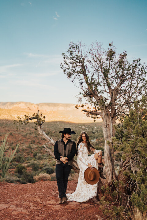 Bride and groom sit on a branch with desert mountain peaks in the background and red dirt after Sedona elopement ceremony