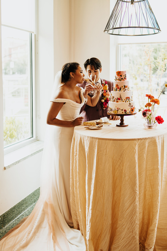 bride and groom sip champagne hand in hand in front of their wedding cake inside of one of the Scottsdale wedding venues. 