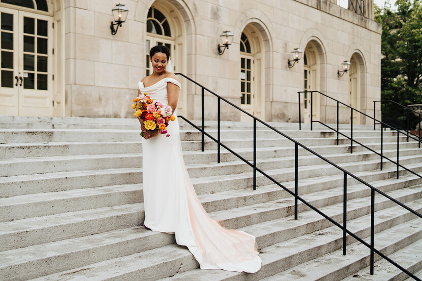 Bride stands at the steps leading up to one of the Scottsdale wedding venues. She is holding a colorful bouquet with pink, yellow, orange, and white flowers. 