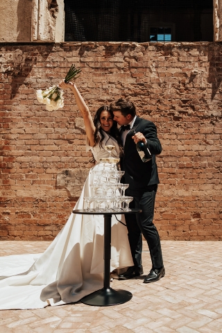 Groom pours champagne onto a tower of champagne glasses while bride holds her bouquet up in the air in excitement at the courtyard of The Abbey on Monroe wedding venue