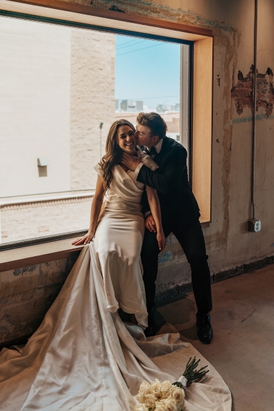 Groom kisses bride on the cheek as she smiles in front of a window in the ballroom of The Abbey on Monroe wedding venue